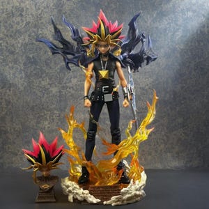 Pode incluir: Uma figura de anime Yami Yugi sobre uma base com chamas amarelas translúcidas, vestindo uma camisa preta sem mangas, calças escuras, um pingente de pirâmide dourada e um acessório de braço prateado, acompanhada por um busto de cabeça separado.