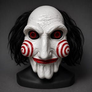 Puede incluir: Una máscara blanca de cabeza completa con un rostro pálido y texturizado, espirales rojas prominentes en las mejillas, una boca pintada de rojo y cuencas oculares oscuras con iris rojos. La máscara incluye cabello negro y descansa sobre una base negra.