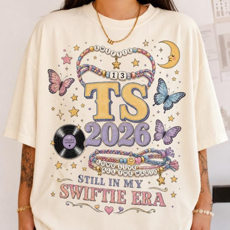 May include: A cream-colored t-shirt featuring a graphic print with the text TS 2026 STILL IN MY SWIFTIE ERA. The design includes illustrations of beaded bracelets, butterflies, a crescent moon, stars, and a vinyl record labeled Taylor's Version.