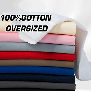 May include: A stack of folded oversized t-shirts in various solid colors including white, cream, light pink, gray, red, brown, blue, navy, and black. The top white shirt features the black text 100% COTTON OVERSIZED printed on the fabric.