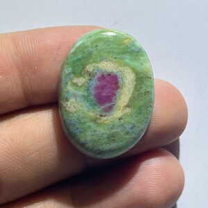 Puede incluir: Una piedra preciosa en cabujón de forma ovalada con una superficie lisa y pulida, que presenta una matriz moteada en verde claro y crema. Una distintiva inclusión de color rosa magenta vibrante se centra en la piedra, rodeada por un halo crema pálido.