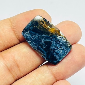 May include: A rectangular polished gemstone featuring a deep blue, swirling, fibrous pattern with a distinct patch of golden-brown mineral inclusions at the top edge. The stone has a smooth, reflective surface and a flat, cabochon-style cut.