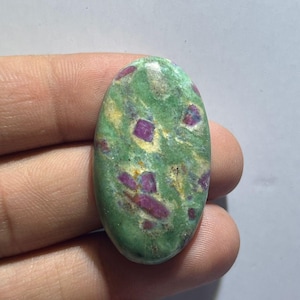 Peut inclure: Une pierre précieuse cabochon de forme ovale présentant une base marbrée verte et jaune clair avec des inclusions de cristaux magenta foncé incrustées sur toute la surface polie, offrant une texture lisse, arrondie et brillante.