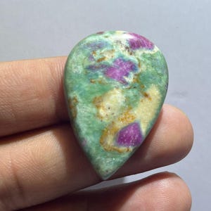 Può includere: Una gemma a cabochon a forma di goccia con una superficie lucida che presenta un motivo maculato di inclusioni magenta brillante su uno sfondo di matrice minerale verde chiaro e giallo crema con sottili venature marroni.