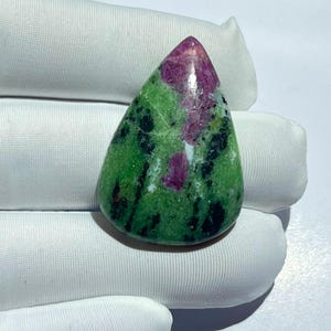 May include: A teardrop-shaped ruby zoisite gemstone featuring a polished surface with a vibrant mix of bright green zoisite, deep magenta ruby inclusions, and dark black hornblende mineral streaks throughout the stone's natural crystalline structure.
