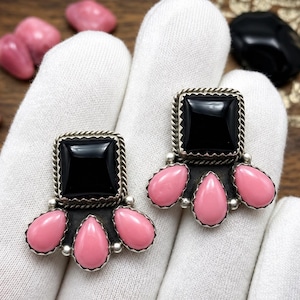May include: A pair of earrings featuring a square black stone set in a twisted metal frame, accented by three teardrop-shaped pink stones arranged in a fan pattern at the base. The metal settings have a dark, oxidized finish around the pink stones.