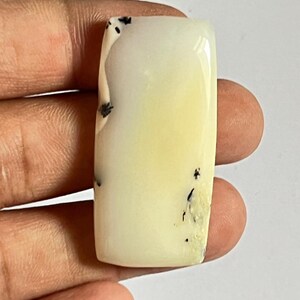 May include: A rectangular, polished cabochon gemstone with a smooth, pale cream-colored surface. The stone features distinct, thin, black dendritic inclusions scattered along its edges and across the face, creating a natural, branching pattern.