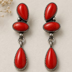 May include: A pair of dangle earrings featuring three red, teardrop and oval-shaped stones set in oxidized silver. Each earring consists of a top teardrop stone, a middle oval stone, and a bottom teardrop stone, all framed by a serrated silver edge.