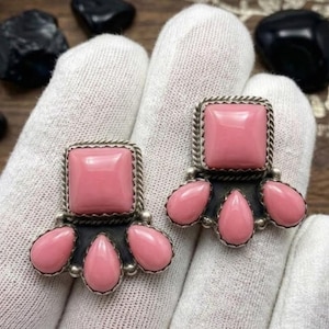 May include: A pair of earrings featuring a square pink stone set in a twisted metal border, positioned above three teardrop-shaped pink stones arranged in a fan pattern. The stones are set in dark metal with a rope-like texture and silver accents.
