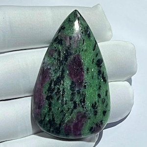Puede incluir: Una piedra preciosa de rubí en zoisita pulida en forma de lágrima, con una base verde vibrante, inclusiones prominentes de cristales de rubí rojo magenta intenso y vetas minerales de hornblenda negra en toda la superficie lisa y brillante.