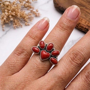 May include: A silver ring featuring a central heart-shaped red stone surrounded by five smaller oval red stones arranged in a paw print pattern. The metal setting is polished silver, and the stones have a smooth, rounded, and opaque finish.