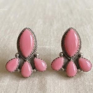 May include: A pair of stud earrings featuring four smooth, pink, teardrop-shaped stones set in silver-toned metal. Each earring has one larger central stone oriented vertically, flanked by three smaller stones at the base, all framed by a twisted rope border.