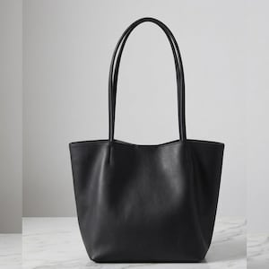 Puede incluir: Un bolso tote negro liso con un acabado mate suave, un dise&ntilde;o de apertura amplia y dos correas de hombro largas y delgadas. El bolso tiene una base estructurada y detalles de costuras verticales donde las correas se unen al cuerpo.