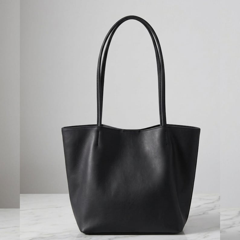 Puede incluir: Un bolso tote negro liso con un acabado mate suave, un dise&ntilde;o de apertura amplia y dos correas de hombro largas y delgadas. El bolso tiene una base estructurada y detalles de costuras verticales donde las correas se unen al cuerpo.
