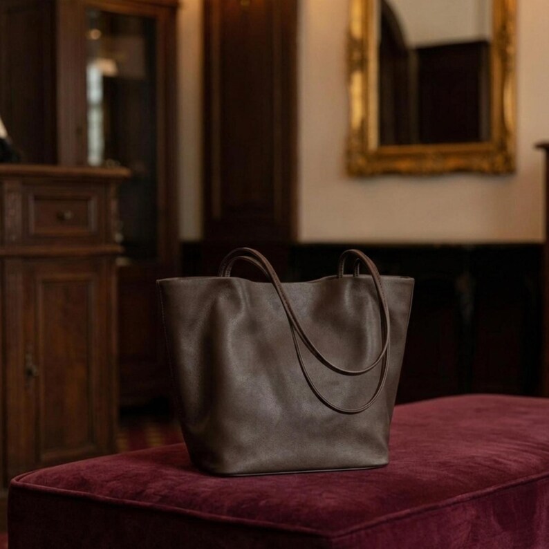 Puede incluir: Un bolso tote de color marr&oacute;n oscuro con un acabado liso y mate, que cuenta con dos asas finas a juego. El bolso presenta un dise&ntilde;o de apertura amplia, una base estructurada y laterales flexibles que crean sutiles pliegues.