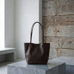 Puede incluir: Un bolso tote de cuero marr&oacute;n oscuro con un acabado liso y mate, una base rectangular ancha y dos correas de hombro largas y delgadas unidas al borde superior. El bolso tiene un dise&ntilde;o sencillo y sin adornos con l&iacute;neas limpias y estructuradas.