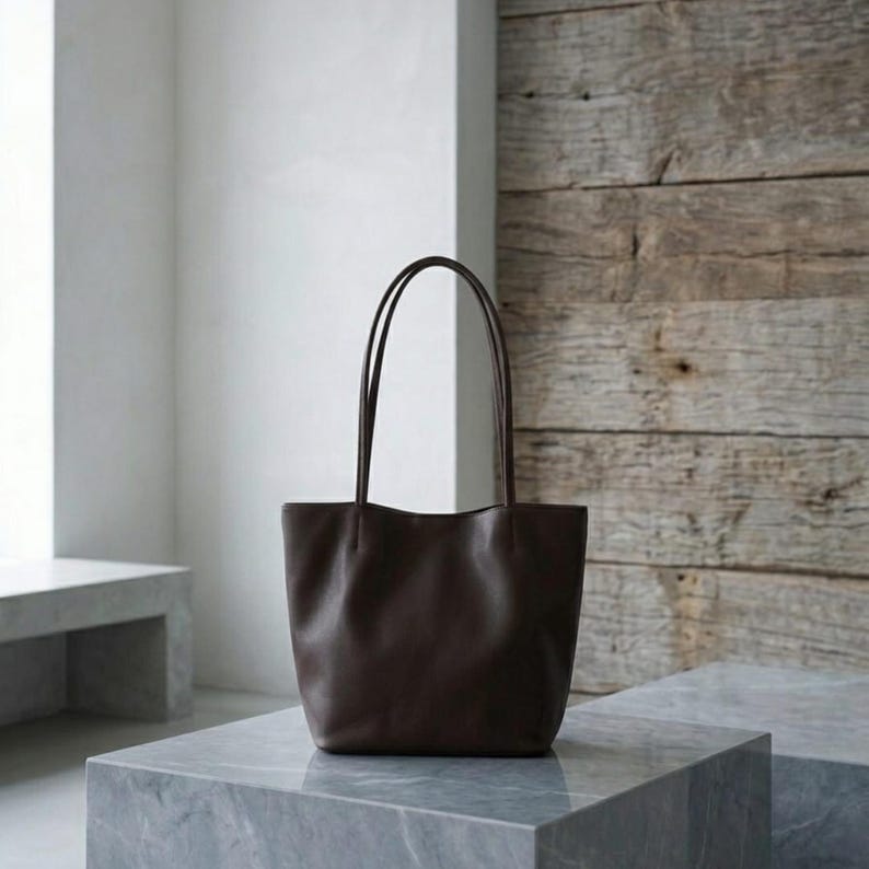 Puede incluir: Un bolso tote de cuero marr&oacute;n oscuro con un acabado liso y mate, una base rectangular ancha y dos correas de hombro largas y delgadas unidas al borde superior. El bolso tiene un dise&ntilde;o sencillo y sin adornos con l&iacute;neas limpias y estructuradas.