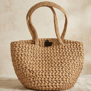 Può includere: Una borsa tote intrecciata color marrone chiaro con un motivo a maglia aperta testurizzato e due manici robusti. La borsa presenta una base arrotondata a secchiello e una piccola linguetta marrone scuro visibile vicino all'apertura superiore.