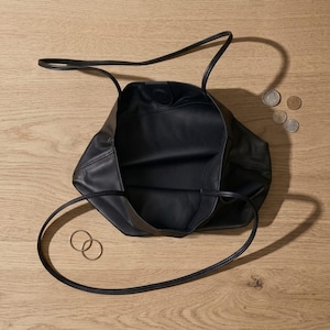 Puede incluir: Un bolso tote de cuero negro con dos asas finas y una abertura superior. El interior est&aacute; forrado en negro, y el bolso se acompa&ntilde;a de dos peque&ntilde;os anillos de metal dorado y tres peque&ntilde;as monedas circulares colocadas cerca en la superficie.