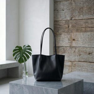 Puede incluir: Un bolso tote negro liso con un acabado mate suave y dos asas de hombro largas y delgadas. El bolso presenta una base estructurada y una abertura superior ligeramente curvada, ofreciendo una apariencia limpia y uniforme sin logotipos.