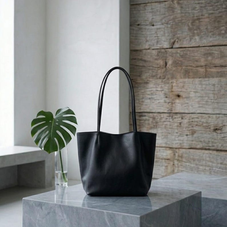 Puede incluir: Un bolso tote negro liso con un acabado mate suave y dos asas de hombro largas y delgadas. El bolso presenta una base estructurada y una abertura superior ligeramente curvada, ofreciendo una apariencia limpia y uniforme sin logotipos.
