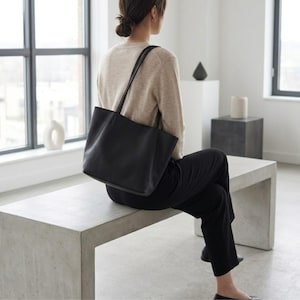 Puede incluir: Un bolso tote de cuero negro con dos correas finas para el hombro, que presenta una base rectangular y un acabado liso y mate. El bolso se lleva sobre el hombro, mostrando su dise&ntilde;o estructurado pero flexible, l&iacute;neas limpias y est&eacute;tica.