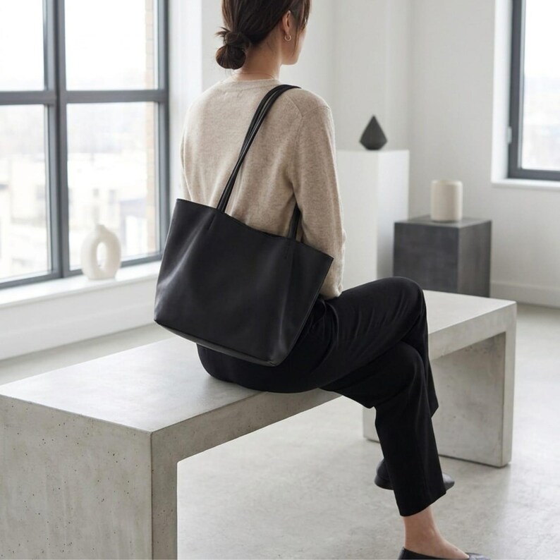 Puede incluir: Un bolso tote de cuero negro con dos correas finas para el hombro, que presenta una base rectangular y un acabado liso y mate. El bolso se lleva sobre el hombro, mostrando su dise&ntilde;o estructurado pero flexible, l&iacute;neas limpias y est&eacute;tica.