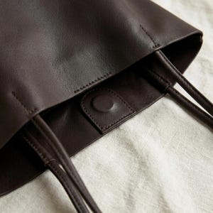 Puede incluir: Un bolso tote de color marr&oacute;n oscuro con un acabado liso y mate, y asas de hombro finas y redondeadas. La abertura interior est&aacute; asegurada con un broche magn&eacute;tico central, con costuras visibles a lo largo del borde superior y la solapa.