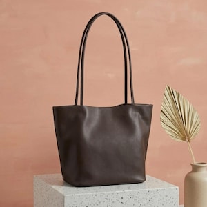 Puede incluir: Un bolso tote de color marr&oacute;n oscuro con un acabado liso y mate, que cuenta con dos asas largas y delgadas. El bolso presenta un dise&ntilde;o de apertura amplia con una base estructurada y l&iacute;neas limpias, mostrando un color s&oacute;lido en el exterior.