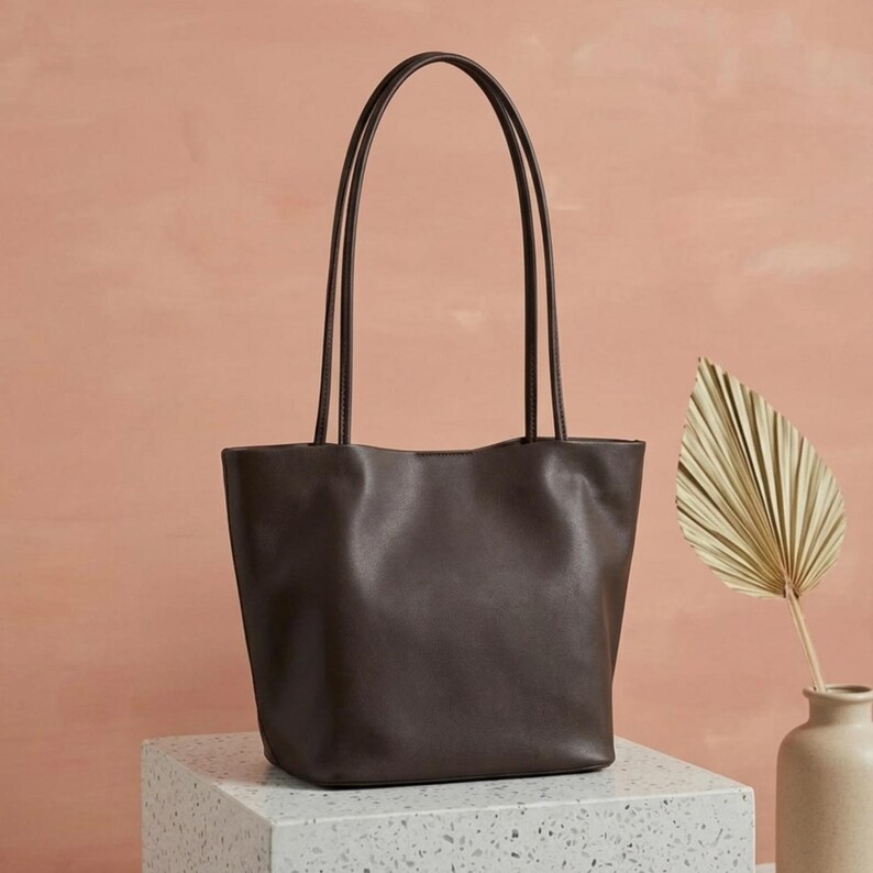 Puede incluir: Un bolso tote de color marr&oacute;n oscuro con un acabado liso y mate, que cuenta con dos asas largas y delgadas. El bolso presenta un dise&ntilde;o de apertura amplia con una base estructurada y l&iacute;neas limpias, mostrando un color s&oacute;lido en el exterior.