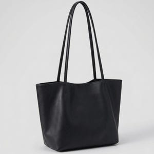 Puede incluir: Un bolso tote negro liso con acabado mate suave, un compartimento principal abierto y dos asas largas y delgadas sujetas a los paneles exteriores superiores con costuras visibles, lo que le otorga una apariencia limpia y estructurada.