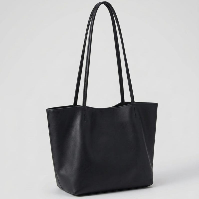 Puede incluir: Un bolso tote negro liso con acabado mate suave, un compartimento principal abierto y dos asas largas y delgadas sujetas a los paneles exteriores superiores con costuras visibles, lo que le otorga una apariencia limpia y estructurada.