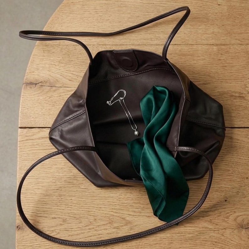 Puede incluir: Un bolso tote de cuero marr&oacute;n oscuro con dos correas finas para el hombro, que contiene un collar plateado con un colgante de perla y un pa&ntilde;uelo de seda verde esmeralda doblado dentro del compartimento principal abierto.