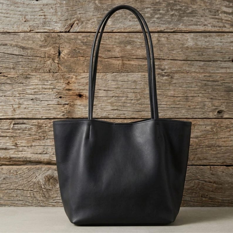 Puede incluir: Un bolso tote negro liso con un acabado mate suave, un dise&ntilde;o de apertura amplia y dos correas de hombro largas y delgadas unidas al borde superior. El bolso tiene una base estructurada y una silueta limpia sin herrajes visibles.