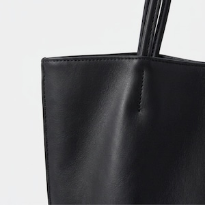 Puede incluir: Un bolso tote negro con acabado liso y mate, costuras visibles a lo largo de los bordes y dos asas de hombro finas y redondeadas unidas al panel superior. El bolso presenta una forma rectangular estructurada con un dise&ntilde;o limpio.