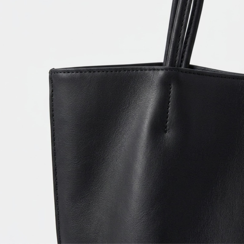 Puede incluir: Un bolso tote negro con acabado liso y mate, costuras visibles a lo largo de los bordes y dos asas de hombro finas y redondeadas unidas al panel superior. El bolso presenta una forma rectangular estructurada con un dise&ntilde;o limpio.