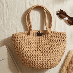 May include: A beige woven tote bag with two matching handles and a rounded base. The bag features a thick, textured braided pattern throughout its construction. A small dark brown rectangular tag is visible near the top center opening of the bag.
