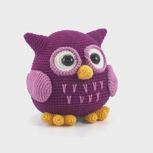 May include: A round, crocheted owl figurine in deep purple yarn. It features large, white-rimmed eyes with black plastic pupils, a small yellow beak, light pink wings, and yellow feet. The chest is decorated with light pink embroidered V-shaped stitches.