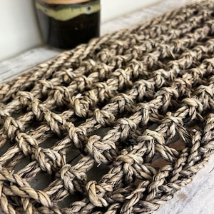 May include: A rectangular mat crafted from thick, natural-colored woven fibers featuring a repeating open-grid knot pattern. The texture is coarse and fibrous, with a light brown and tan color palette throughout the interlocking rope-like strands.