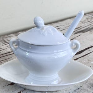 May include: A light blue ceramic sugar bowl with a matching lid and a small spoon. The bowl features two side handles and sits on a matching oval saucer. The lid has a small knob handle and a subtle embossed leaf pattern on the top surface.