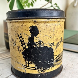 May include: A cylindrical metal tin with a distressed yellow finish and a black lid. The exterior features a black silhouette graphic of a person sitting in a rocking chair while knitting, with decorative line work extending across the surface.