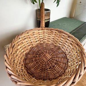 May include: A round, light brown woven wicker basket featuring a single upright handle attached to the rim. The basket has a circular base with a square-shaped center weave pattern and a thick, braided border around the top edge of the structure.