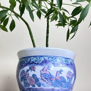 May include: A round ceramic planter featuring a white base with a hand-painted design in shades of blue, purple, and pink. The decorative pattern includes stylized peacocks, floral motifs, and geometric borders. A green plant grows from the top.