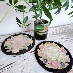 May include: Two round punch needle coasters featuring a central cream-colored circle surrounded by a floral wreath pattern in shades of pink, blue, green, and yellow, all set against a dark brown border with a textured, looped yarn finish.