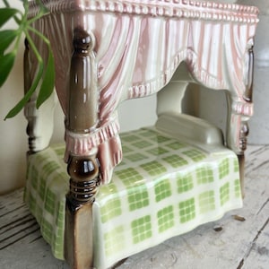 May include: A ceramic four-poster bed figurine featuring brown carved posts, a pink canopy with draped fabric details, and a green and white checkered quilt. The piece includes a raised headboard and a glossy finish on all surfaces.