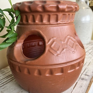 May include: A terracotta-colored strawberry planter with a wide, circular opening at the top and multiple side pockets for planting. The exterior features a molded geometric pattern of diamonds and horizontal bands around the rim and base.