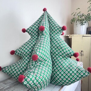 May include: A three-dimensional, tree-shaped fabric pillow featuring a green background with a repeating pattern of small white and red floral motifs. The points of the tree are decorated with small, round, red pom-poms attached to the tips.
