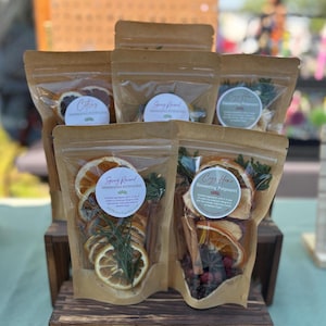 Könnte beinhalten: Mehrere transparente Beutel gefüllt mit getrockneten Früchten und Kräutern, beschriftet mit "Citrus", "Spring Renewal" und "Cozy Home". Die Beutel sind auf einem Holzständer angeordnet. Jeder Beutel enthält eine andere Mischung von Zutaten für Potpourri zum Köcheln.