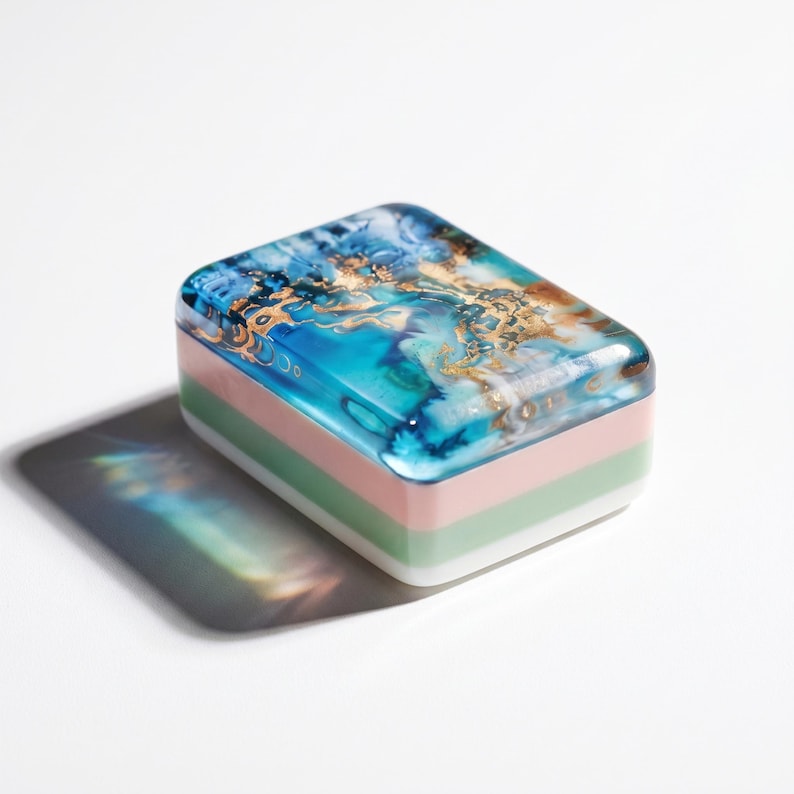 May include: A rectangular block with a glossy top surface featuring a swirling pattern of blue, teal, and gold metallic accents. The sides of the block consist of three distinct horizontal layers in shades of light pink, sage green, and white.