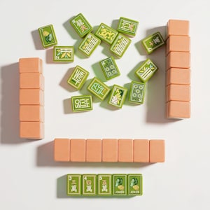 May include: A set of mahjong tiles including several peach-colored blank tiles arranged in stacks and rows, alongside green tiles featuring various symbols, numbers, Chinese characters, and the word JOKER printed in white, black, and gold ink.